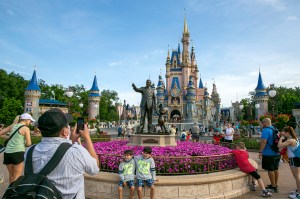 Several park visitors take photos of two kids standing in front of a statue of Walt Disney with a castle in the background.
