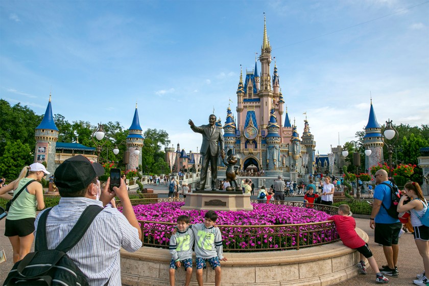 Several park visitors take photos of two kids standing in front of a statue of Walt Disney with a castle in the background.