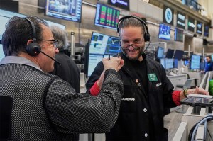 Two options traders wearing headsets fist pump each other on the floor of the New York Stock Exchange, surrounded by trading screens displaying market data.