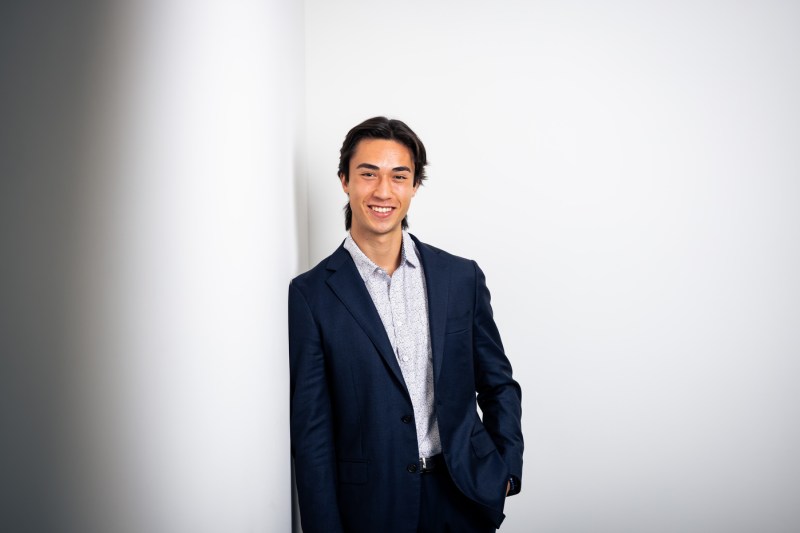 Portrait of Cameron Little wearing a blue and white patterned shirt under a blue blazer. Little is leaning up against the corner of two white walls. 