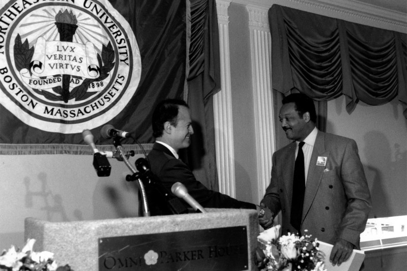 A black-and-white photo of Jesse Jackson shaking hands with a man at a podium and the Northeastern crest hanging on the wall behind them.