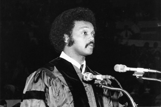 A black-and-white photo of Jesse Jackson, in commencement regalia, standing before microphones.