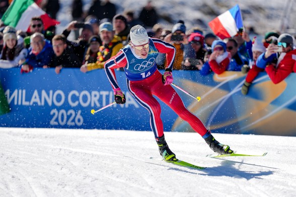 Johannes Hoesflot Klaebo wearing a blue and red ski suit cross country skis across the snow in front of a crowd at the 2026 Winter Olympics.