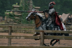 An armored knight with a red-plumed helmet rides a brown horse at speed while holding a jousting lance, captured in motion past wooden fencing in a medieval tournament arena.