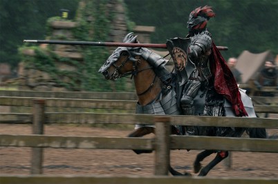 An armored knight with a red-plumed helmet rides a brown horse at speed while holding a jousting lance, captured in motion past wooden fencing in a medieval tournament arena.