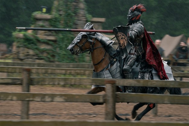 An armored knight with a red-plumed helmet rides a brown horse at speed while holding a jousting lance, captured in motion past wooden fencing in a medieval tournament arena.