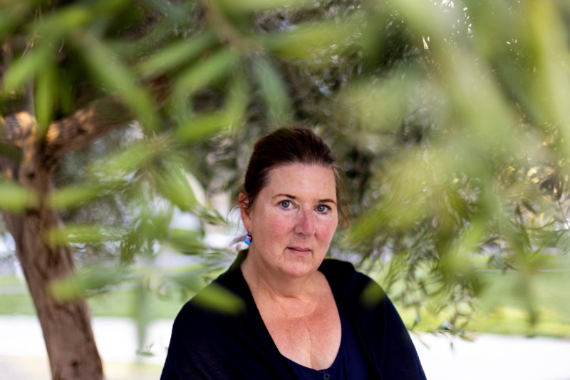 Kathryn Reiss, wearing a black v-neck top, photographed outdoors through soft-focus green foliage that frames the portrait, with natural leafy background and dappled lighting.