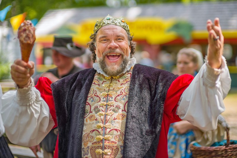Kirk Simpson smiles with outstretched arms in colorful medieval costume at an outdoor Renaissance faire event, with other costumed participants visible in the blurred background.