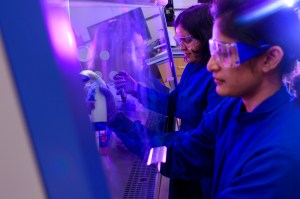 Two female students in blue lab coats and protective eyewear stand in front of a glove box, conducting an experiment at a lab.