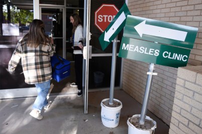 A person wearing jeans and a plaid jacket entering a building through a door propped open. Next to the door is a green sign that says 'Measles Clinic' with an arrow pointing inside the building.
