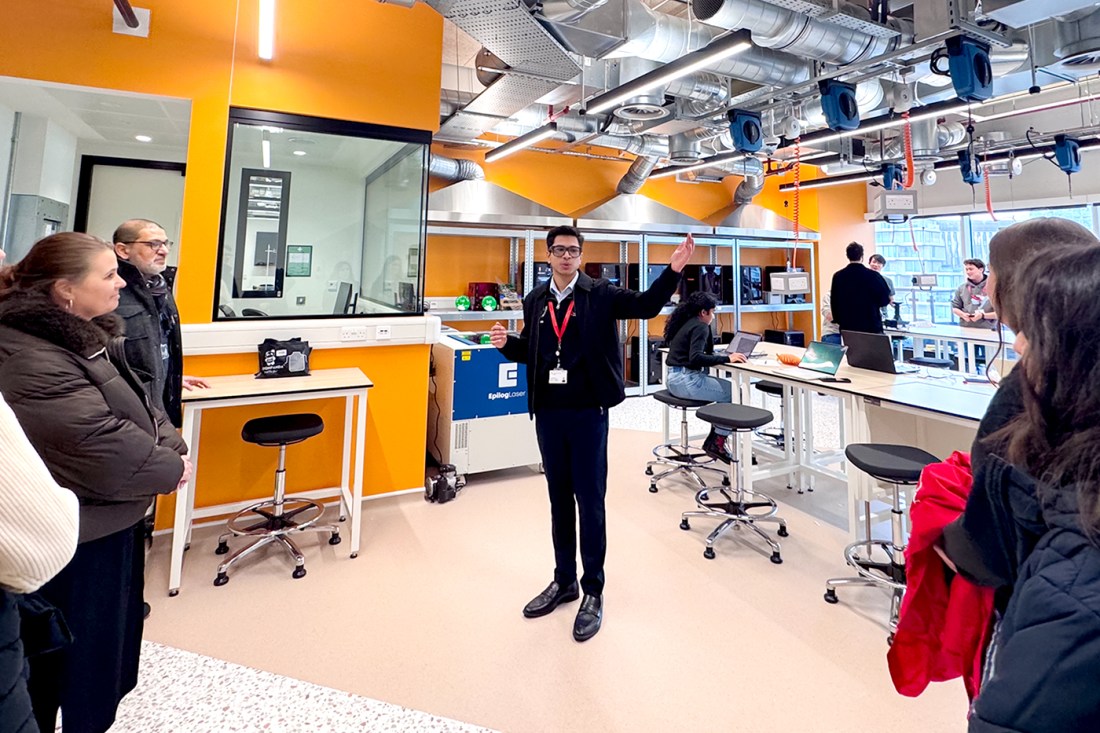 A tour group consisting of four people and a tour guide stands inside a lab space with orange walls long desks pushed together in a two lines and stools and an industrial ceiling with exposed pipes.