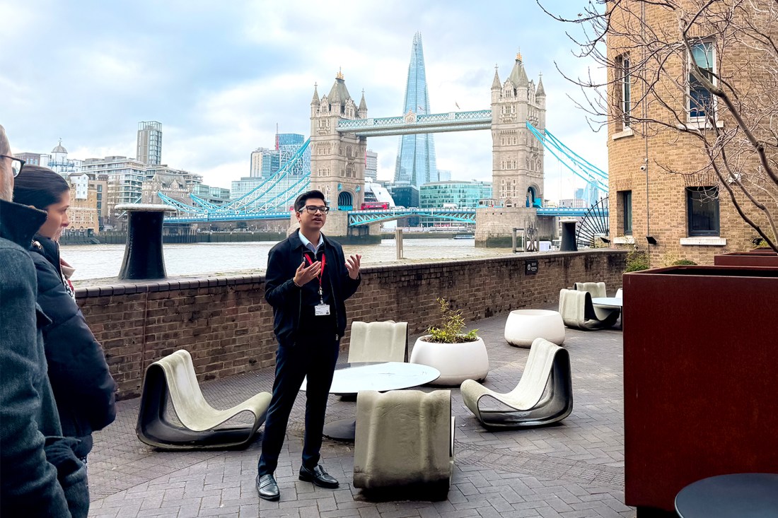 A male tour guide wearing black glasses, black jacket, black pants and black shoes, speaks to a tour group outside by the Thames with the Tower Bridge in the background.