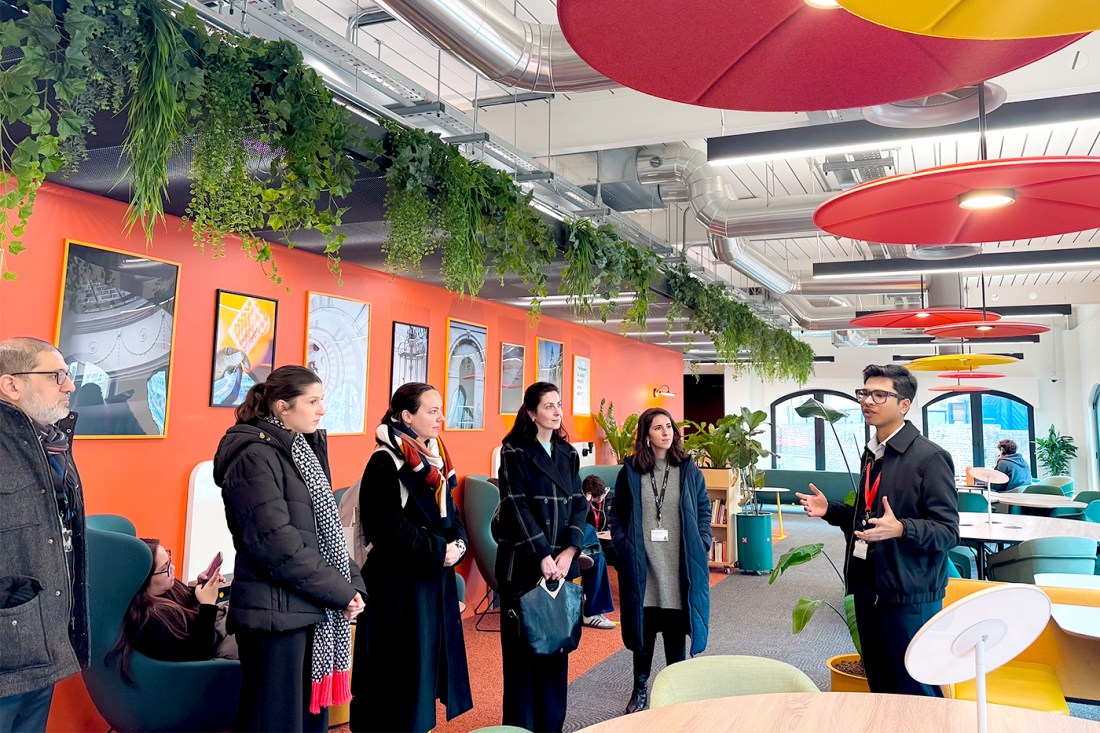 A group of five people stands in front of a male guide in a big common space with tables and armchairs. An orange accent wall is behind them adorned with photographs, green plants hanging above it and red saucer plate light fixtures coming down from the ceiling.