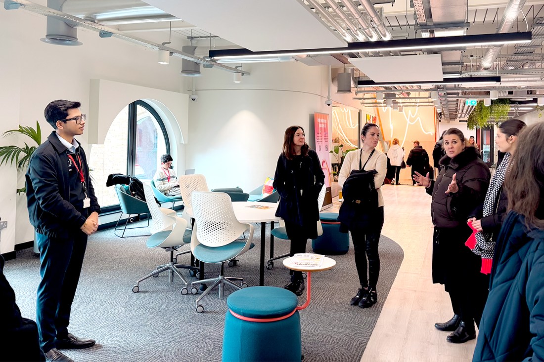 Tour participants stand in a common space with an industrial ceiling, white walls and tables and chairs spread out for work.