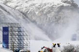 Snow cannons shoot snow up a steep incline in a mountainous area of the Milano Cortina 2026 Winter Olympics. Scaffolding with the Olympic rings stands nearby.