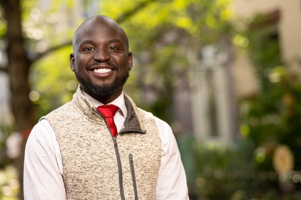 A bald Black man with a short beard smiles while wearing a vest over a suit and red tie.