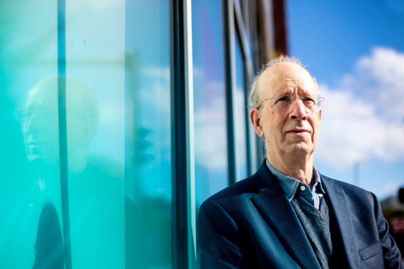 Phil Brown, wearing glasses and a dark suit, in an outdoor portrait against a modern glass building with turquoise reflective panels and blue sky.