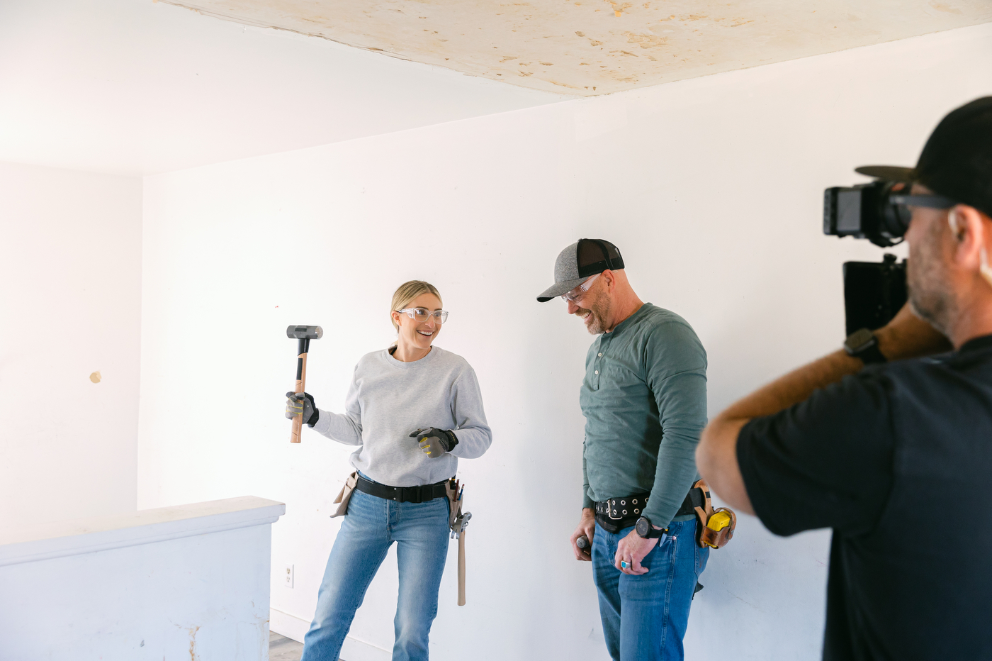 Jasmine Roth and another person both wear tool belts and protective goggles while a camera crew-member shoots.