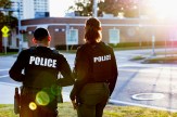 Two police officers standing on a street corner with their backs to the camera. On the back of their black uniforms the word 'POLICE' is in all white. They are lit by the glow of the setting sun.