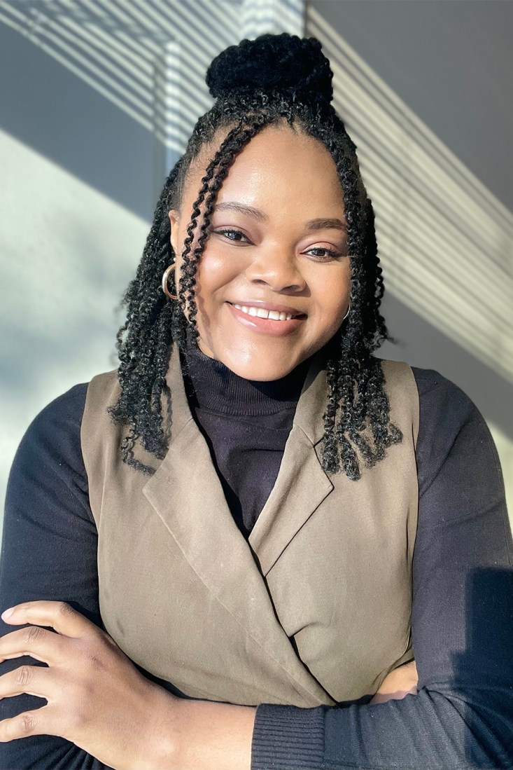Portrait of a woman wearing a taupe vest over a black turtleneck, with gold hoop earrings and her hair pulled into a half up half down bun.