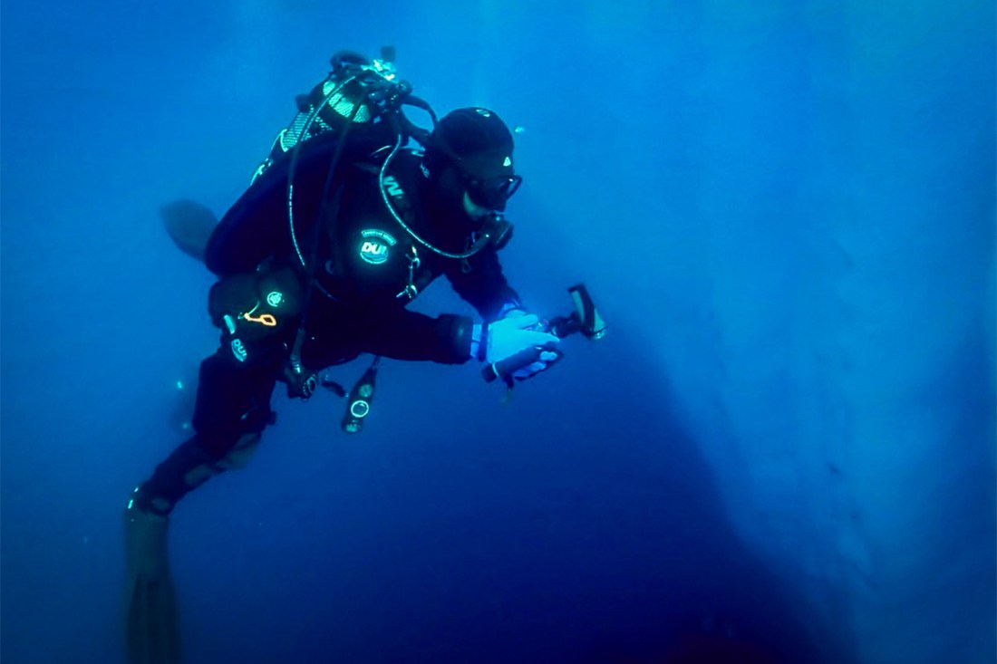 A scuba diver underwater wearing full gear and holding a light, swimming through deep blue water.