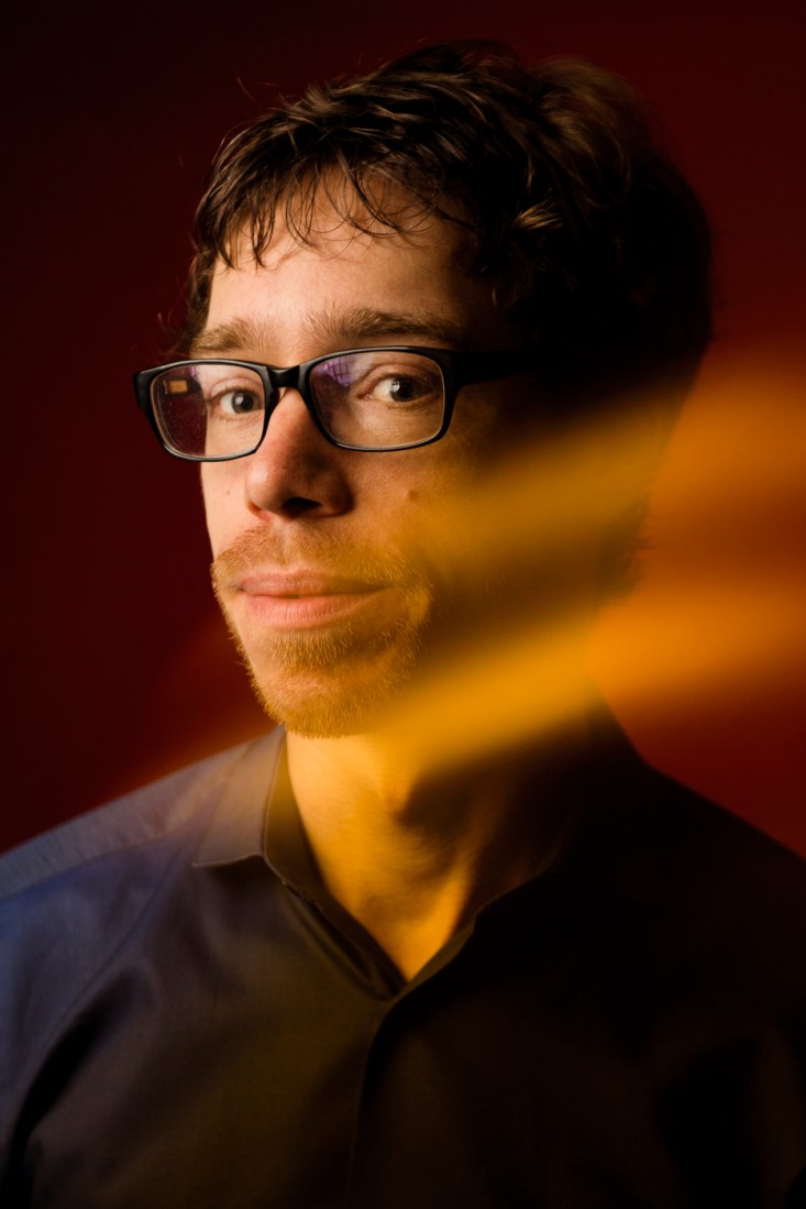 Portrait of Chris Riedl wearing a dark shit, lit with warm golden red light against a deep red background.