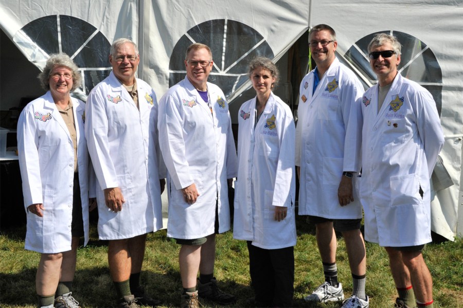 A group of people in white lab coats with a "STEM Quest" embroidered on them pose for a photo in front of a white events tent.