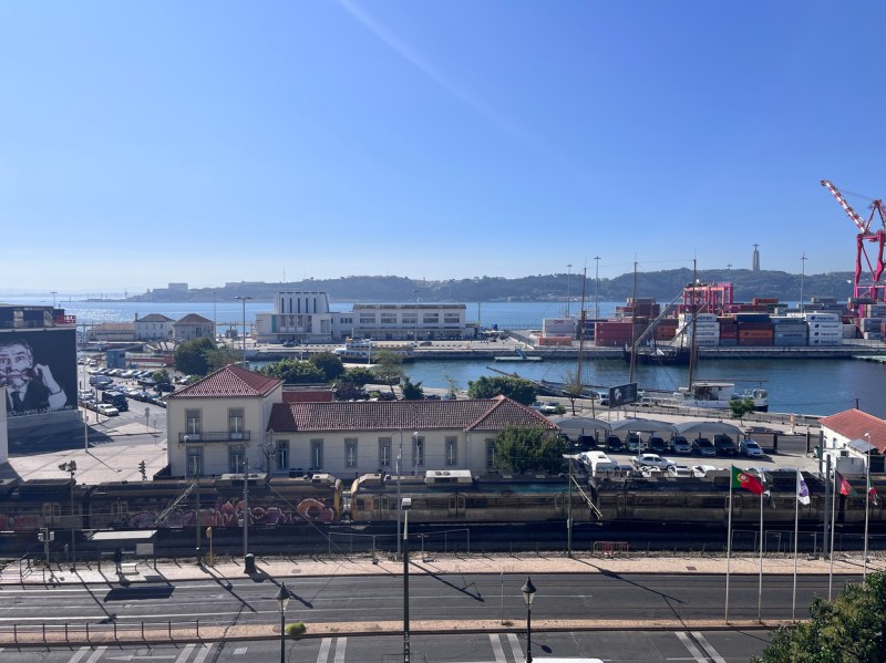A view of a working harbor with cranes, docked cargo ships, and waterfront buildings under a clear blue sky. 