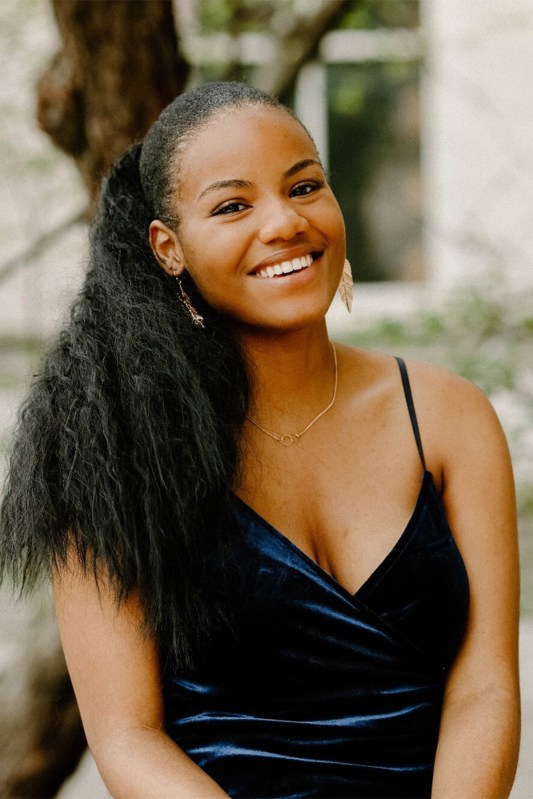Portrait of a woman wearing a blue velvet dress, dainty necklace, and dangly earrings with her hair up in a ponytail.