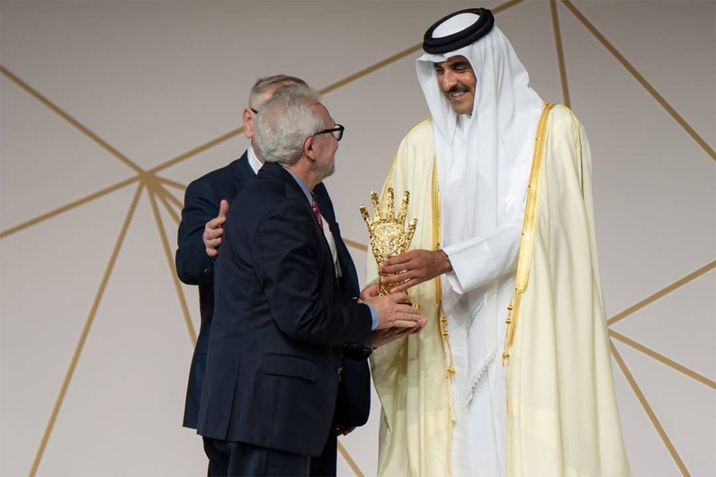 Nikos Passas, wearing a dark suit, received a golden trophy in the shape of a hand, from a man wearing traditional Qatari robes. and head covering. They are standing in front of geometric wall panels. 