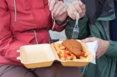 Someone in a red jacket shares a to-go container with someone in a green jacket. Each holds a plastic fork above a fish and chips meal.