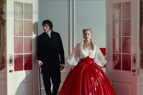Jacob Elordi wears a nineteenth century suit next to Margot Robbie, who wears a bright red skirt and white top. They stand in an open doorway with sky behind them.
