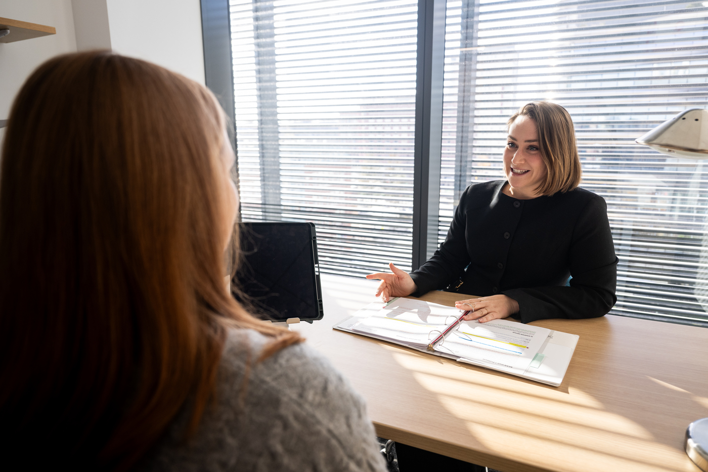 A woman sits in front of a window at a desk, talking to another person, in foreground, with long hair.