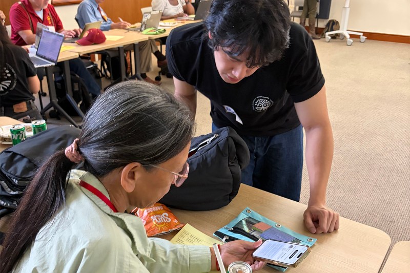 An instructor in a black shirt leans over to assist participants working at tables with laptops and materials.