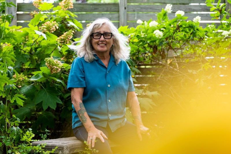 Kathleen Coyne Kelly sits outdoors surrounded by lush green garden foliage and yellow flowers in a residential backyard setting with soft natural lighting.