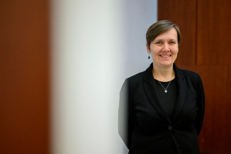 A woman wearing a black top and gold necklace stands against a wooden wall.