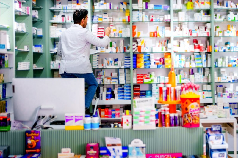 A person in a white pharmacist's coat stands on a small ladder to pull a bottle off a crowded shelf of medicine bottles in a pharmacy environment.
