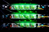Close-up of stacked network servers with rows of ports glowing green and multiple ethernet cables connected, viewed from the front.