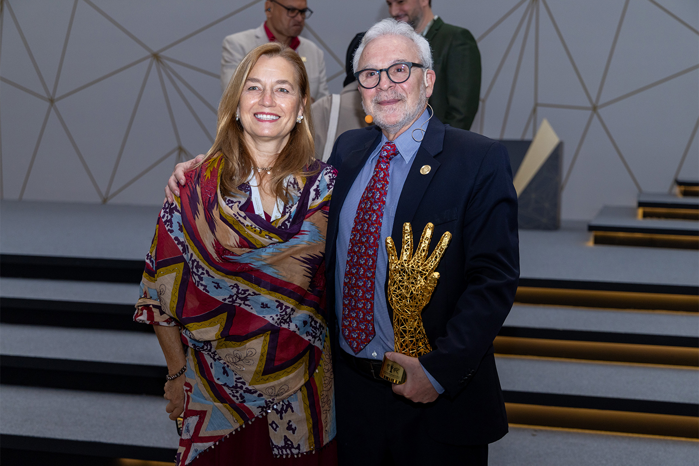 Nikos Passas wearing glasses, a dark suit, a blue shirt and a red patterned tie, holding a gold trophy in the shape of a hand. He is standing next to and with his arm around a woman with shoulder length blonde hair wearing a colorful patterned shawl. 