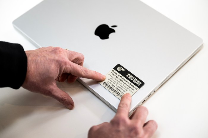 A closeup image of hands putting a registration sticker on the front right corner of a silver Apple laptop on a white table.