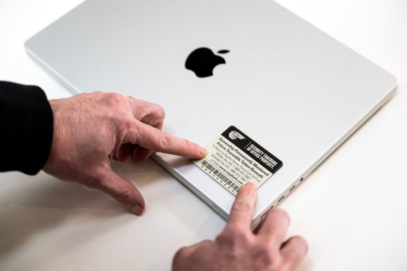 A closeup image of hands putting a registration sticker on the front right corner of a silver Apple laptop on a white table.