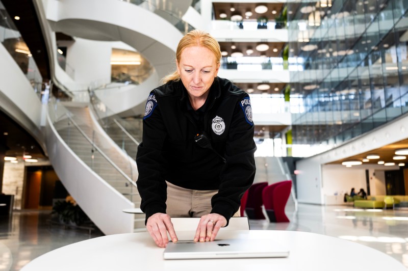 A woman in a black police jacket and light slacks puts a registration sticker on a laptop resting on a table in a office building full of windows and glass.