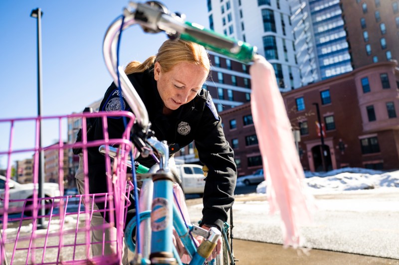 A woman in a black police jacket and light slacks puts a registration sticker on a blue bike with a pink basket and a green handlebar with a light pink tassel outside on a sunny day.  One can see sidewalks, snow, cars and residential multiunit buildings in the background.  
