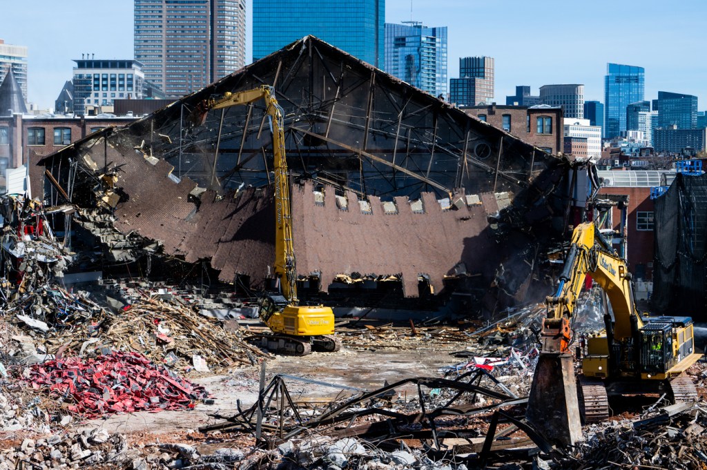A site of the Matthews Arena deconstruction on a sunny day with the city skyline behind the arena's slumped roof and machinery working among the rubble in the foreground.