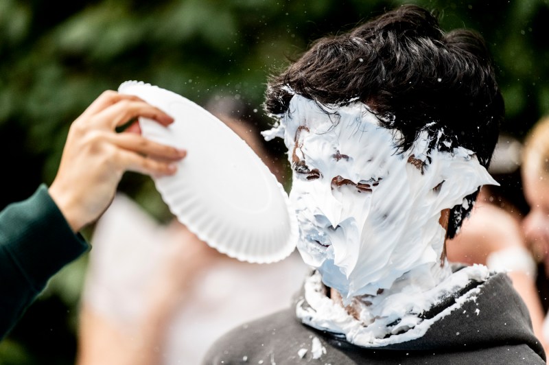 A close up of a student receiving a pie to the face. Whipped cream covers their faces, hair and ears.