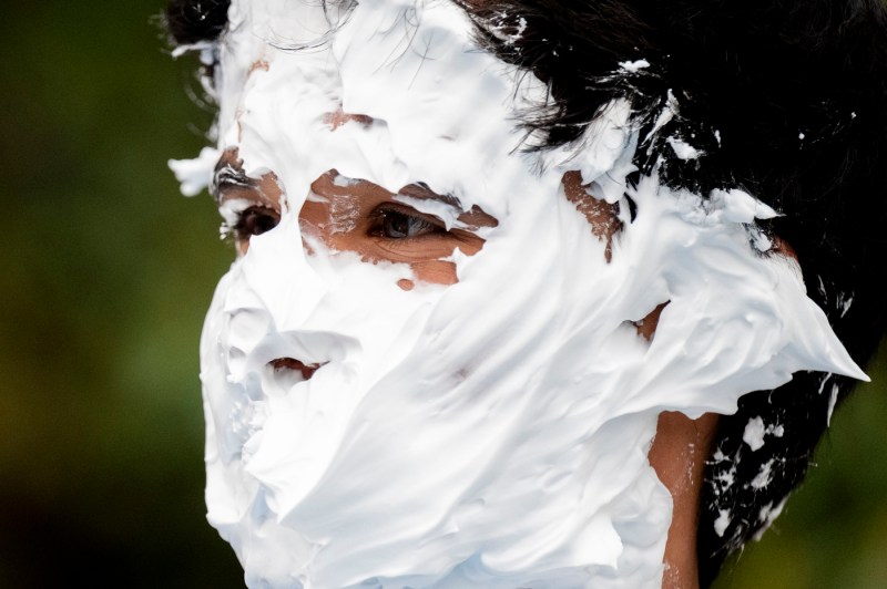 A close up of a student receiving a pie to the face. Whipped cream covers their faces, hair and ears.