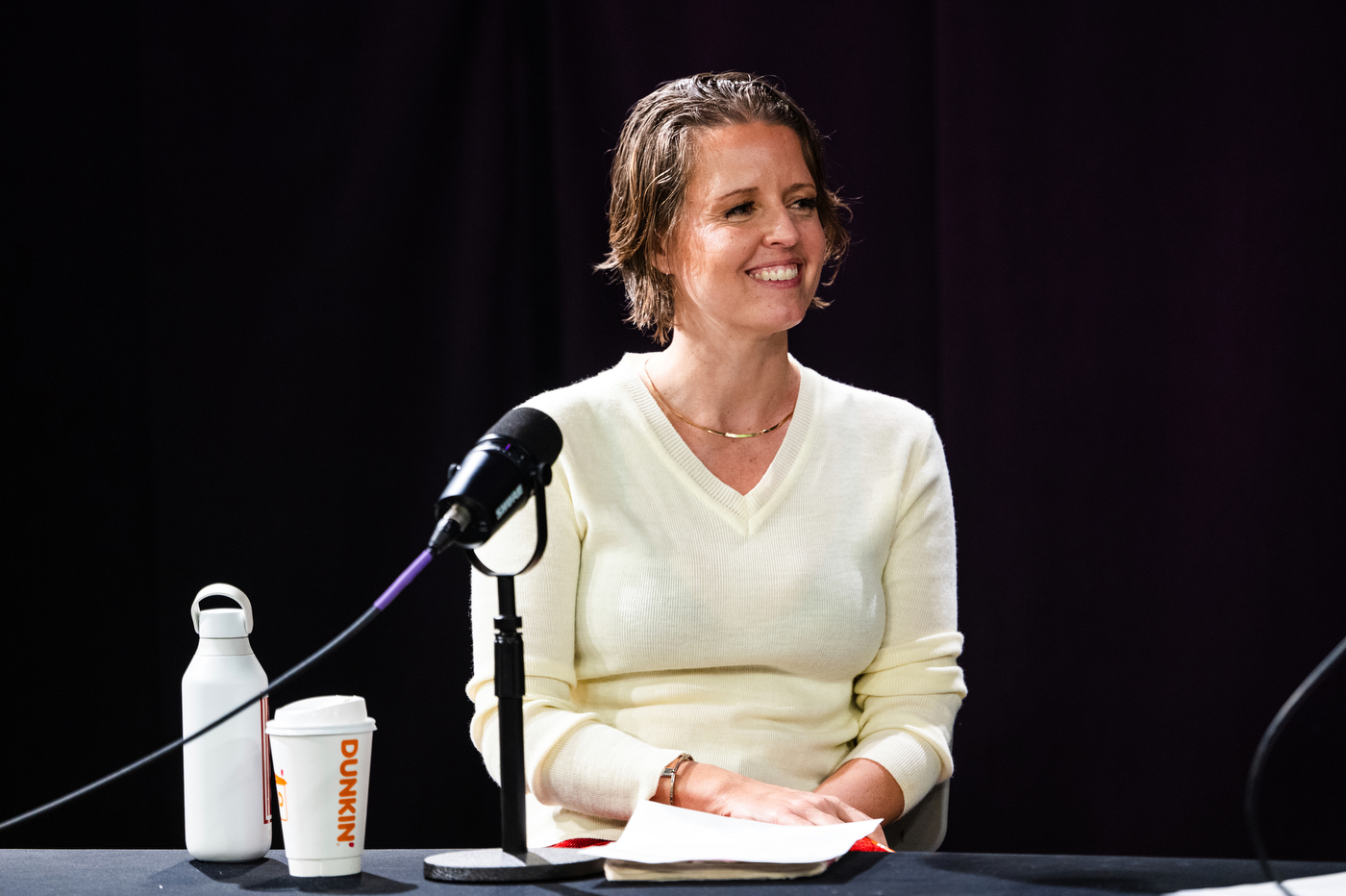 Emily White wearing a cream sweater smiles while seated at a desk in front of amicrophone, with a Dunkin cup and a water bottle sitting next to each other. 
