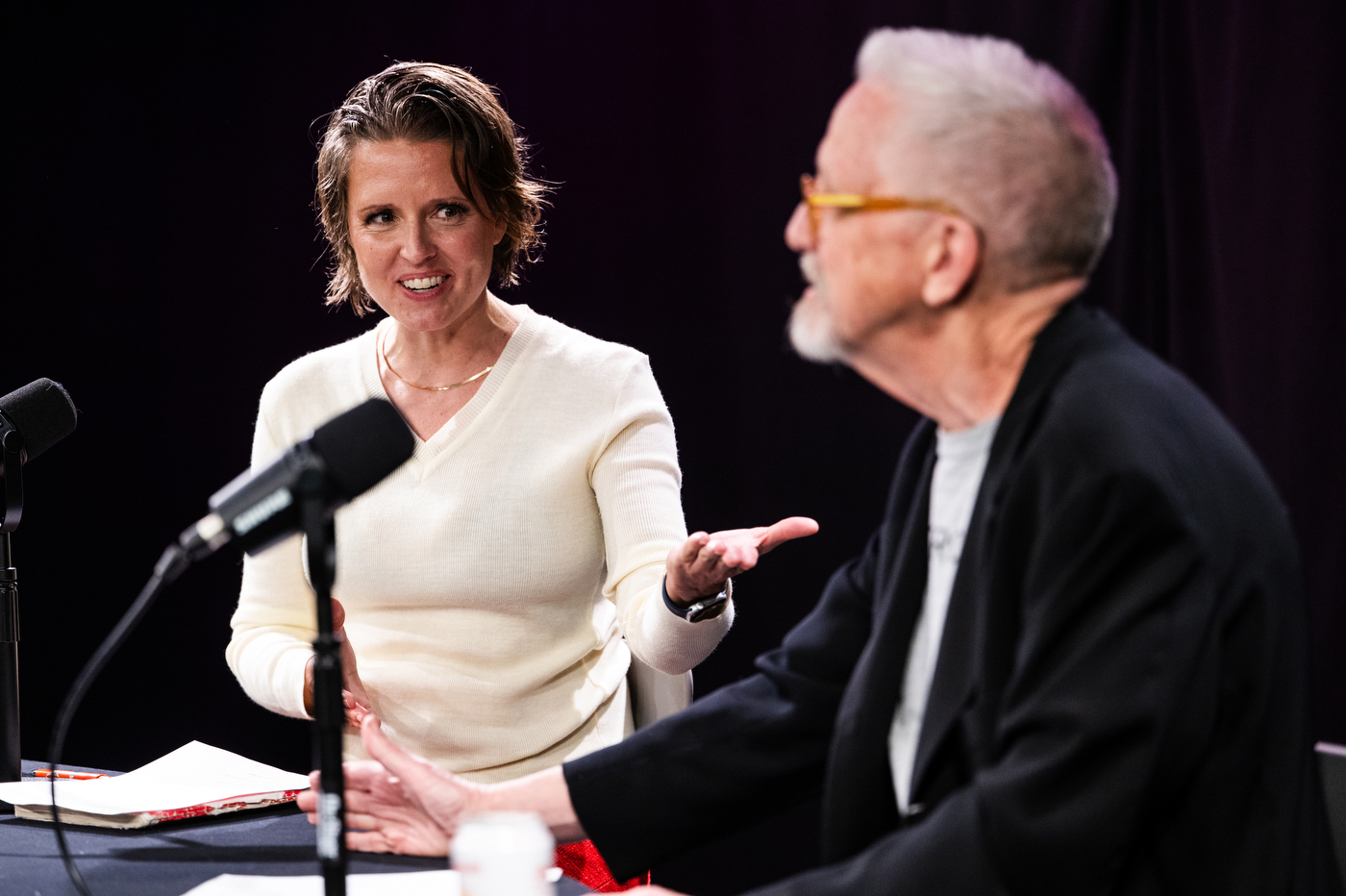 Emily White in a cream sweater gestures with one hand to David Herlihy while speaking into a microphone. 