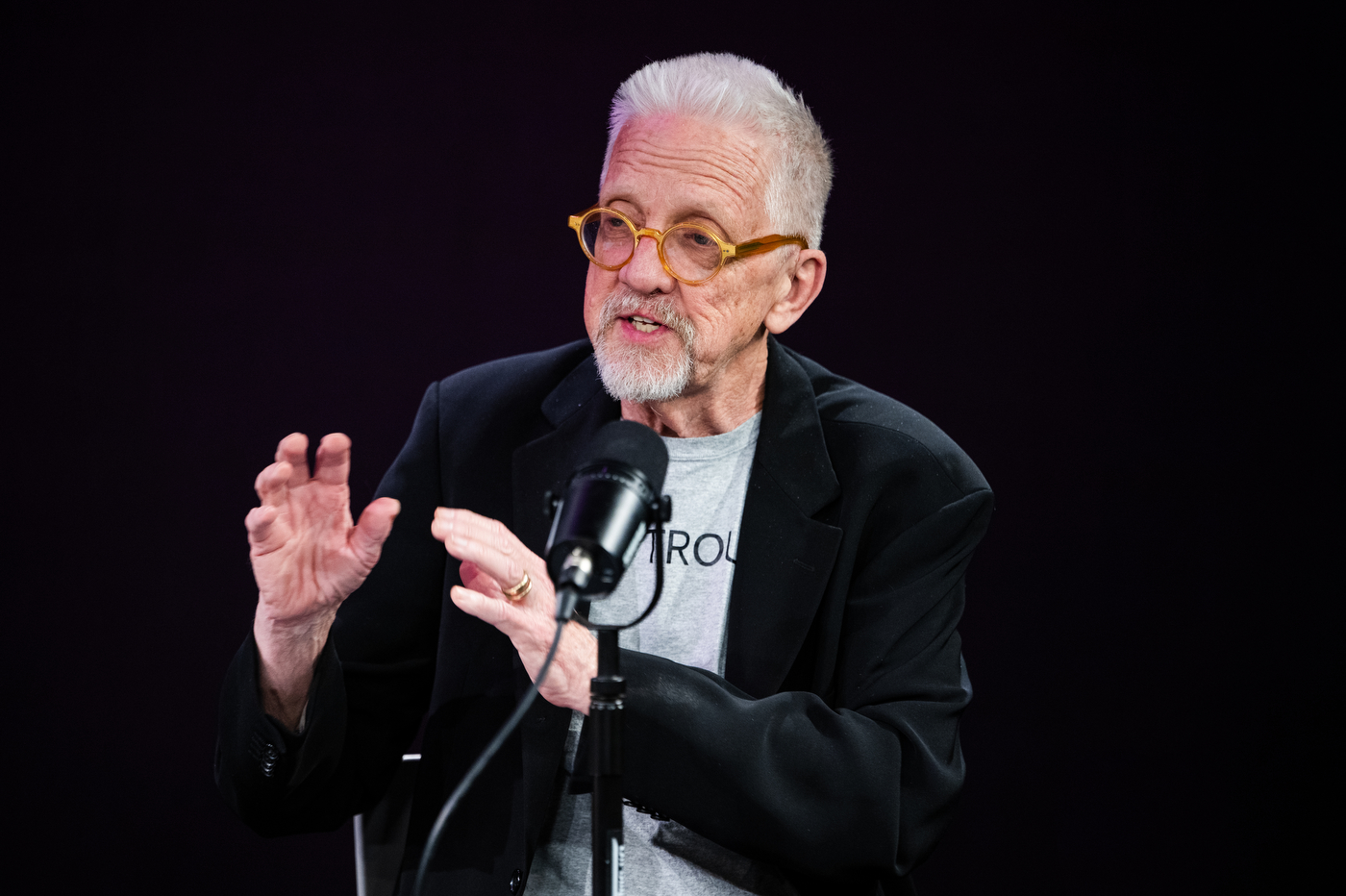 David Herlihy wearing orange framed glasses speaks animatedly with his hands raised into a microphone, wearing a black blazer over a gray t-shirt. 