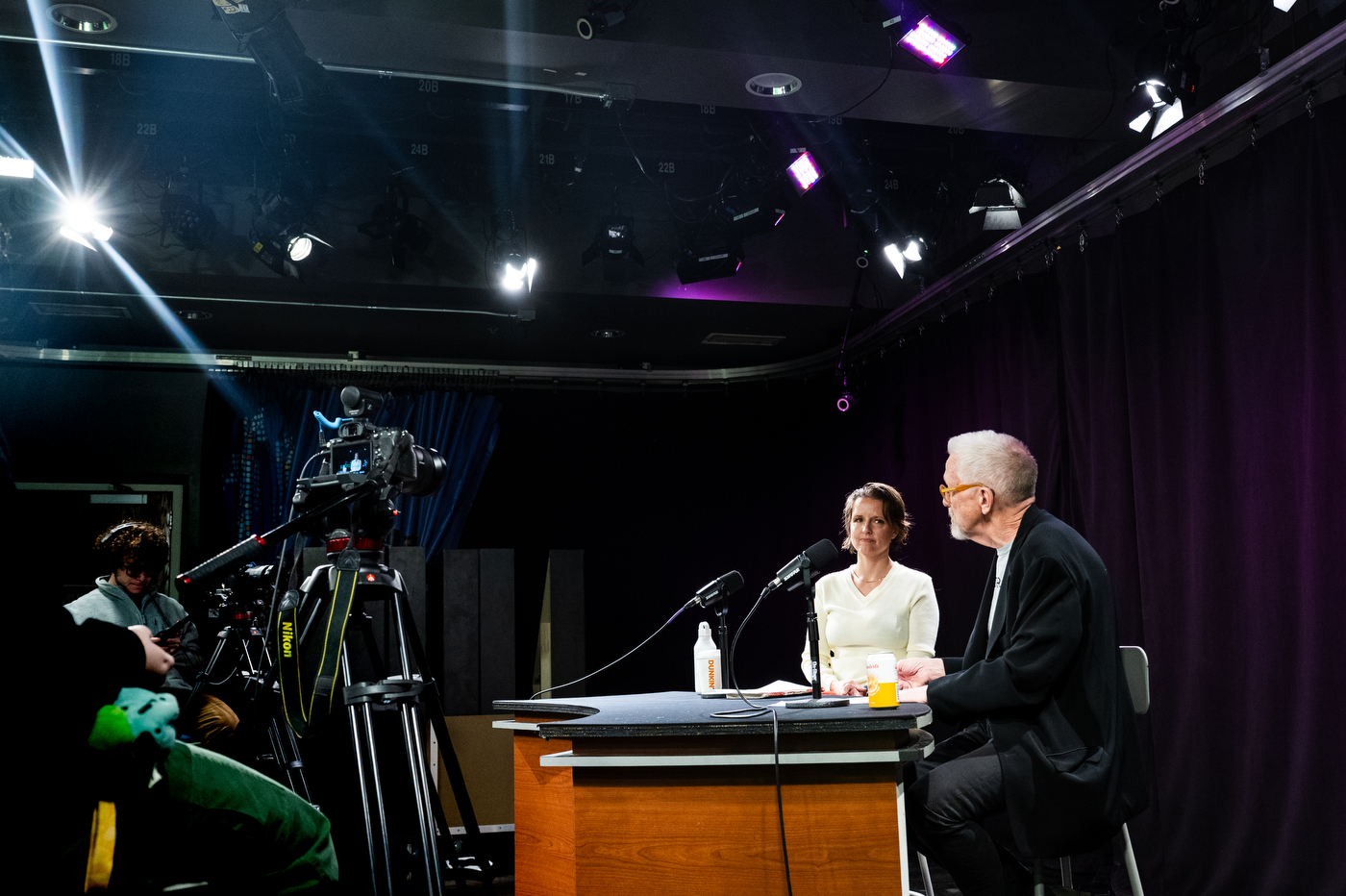 A wide shot of a podcast studio showing Emily White and David Herlihy seated at a wooden desk under bright lights in front of microphones. Camera operators and equipment is visible in front of the desk. 